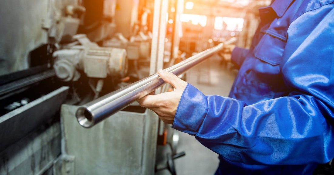 A worker in blue coveralls holds up a metal pipe in front of metal fabrication equipment. The sun is streaming in from above.