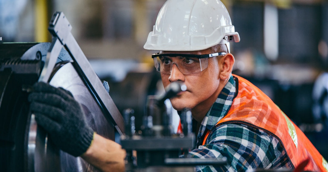 A man with a hardhat and safety goggles is inspecting a metalworking saw. He is carefully checking the condition of the blade.