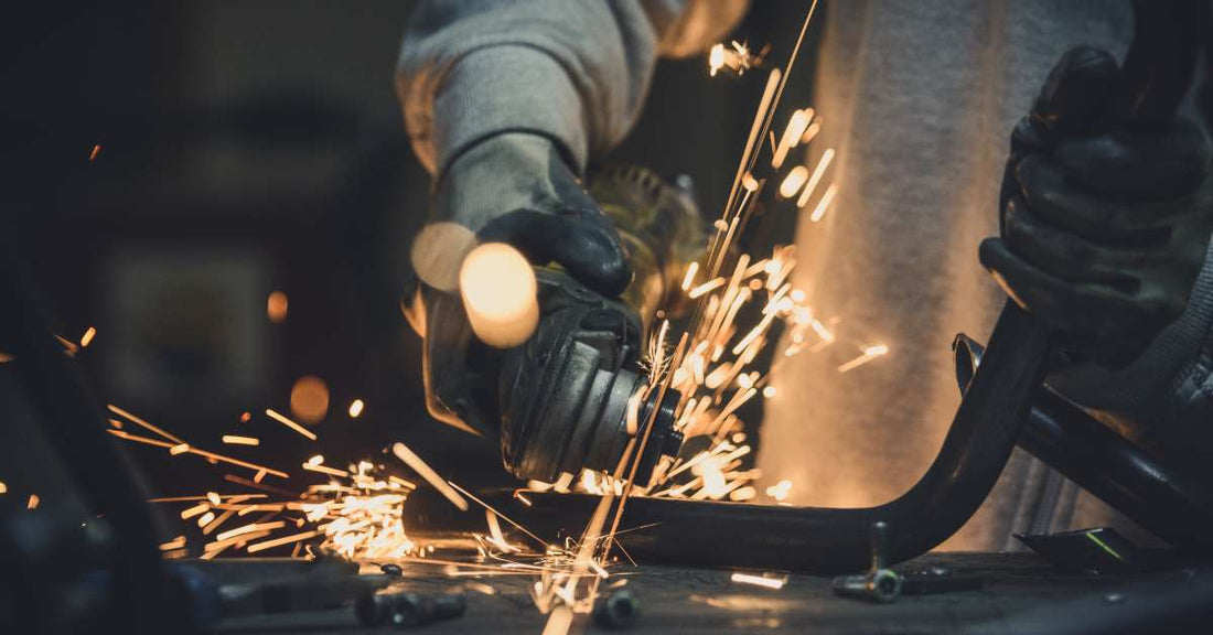 A man in his metalworking shop is cutting a metal pipe at an angle with a handsaw. Sparks are flying from the metal blade.