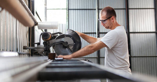 A man in a white t-shirt and protective glasses holds a circular saw and a metal workpiece to make a cut.