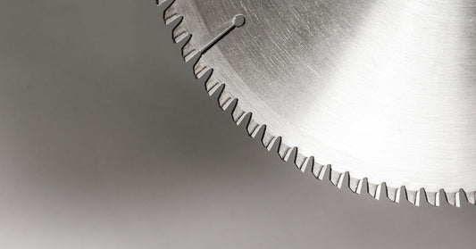 A close-up of part of a round metal saw blade with tipped edges in front of a plain gray background.
