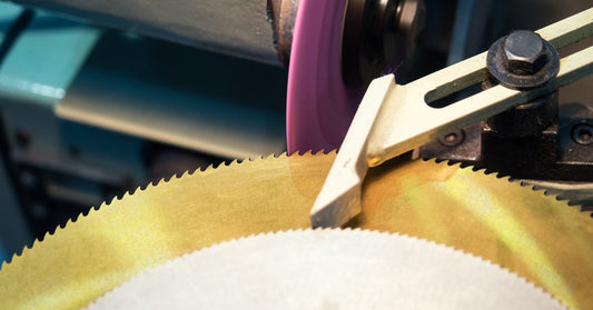 A silver saw blade sits on top of a gold saw blade and is grinded by a piece of metal attached to machinery.