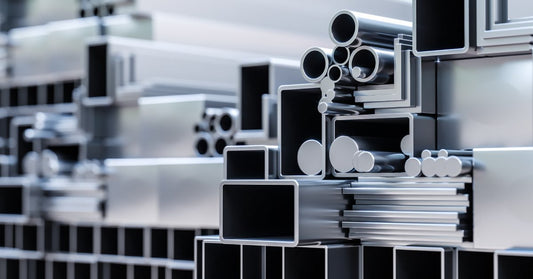 A variety of different types and sizes of long metal pieces sit stacked together on a shelf, reflecting light.