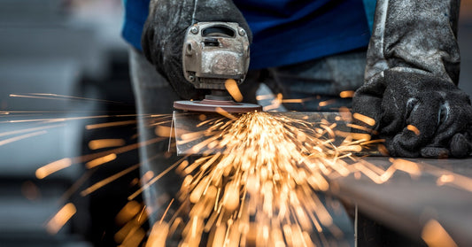 An industrial worker uses an angle grinding saw to cut metal. Sparks are flying off of the piece of metal.