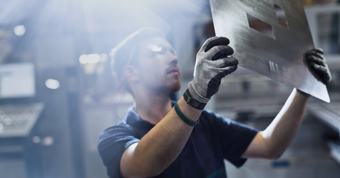  An industrial worker wearing safety gloves holding up a sheet of steel that has rectangular holes punched out of it.