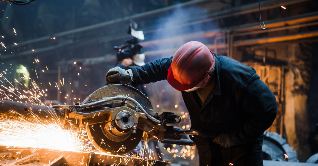 An industrial worker in a hard hat and coveralls using a large saw to cut metal, sending sparks flying into the air.