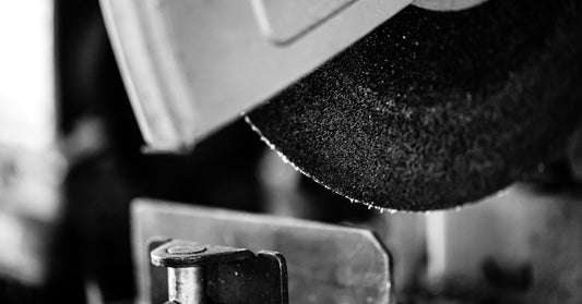 A black abrasive chop saw blade is at close range. It is inside of a machine and hovers above a piece of metal.