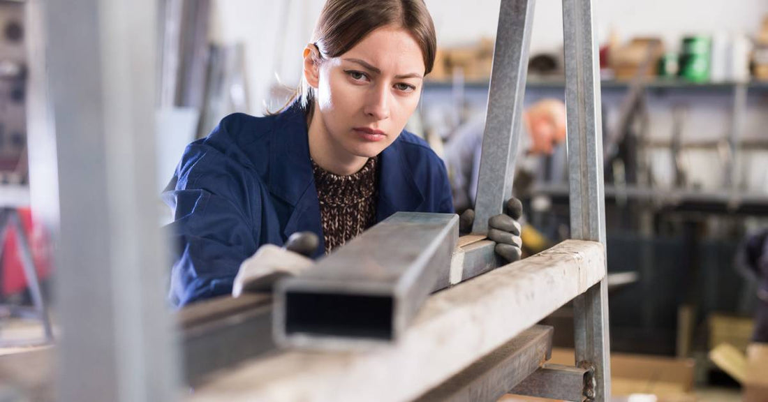 A woman in a blue jacket and gloves looks down on a piece of cut metal in a metalworking shop.