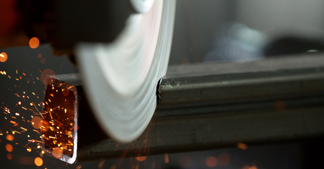 A spinning circular saw blade is cutting through a rectangular piece of metal. Hot red sparks are flying off to the side.