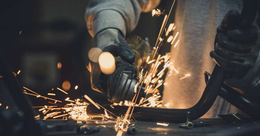 A man in his metalworking shop is cutting a metal pipe at an angle with a handsaw. Sparks are flying from the metal blade.