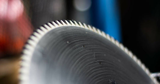 A spinning aluminum saw blade with carbide-tipped teeth shown against a blurred background.