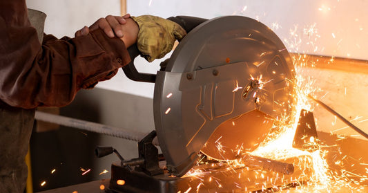 A man wearing protective gloves is using a metalworking saw. The blade is cutting through metal, sending sparks into the air.